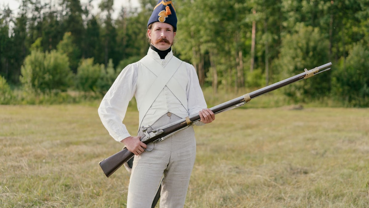 Man dressed in historical uniform with musket in a field setting.